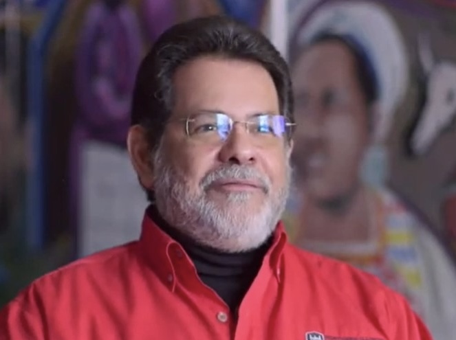Portrait of Luis Santos-Rivas in a red shirt, with a colorful mural behind him